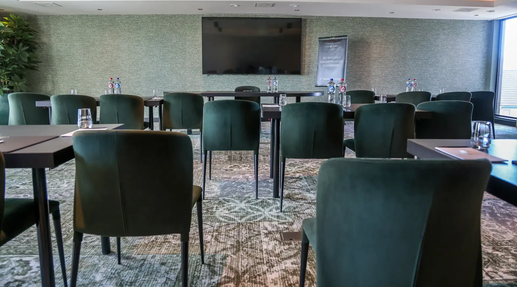 Konferenzraum mit einer Schulaufstellung mit braunen Tischen mit Gläsern und Wasser und grünen Stühlen.