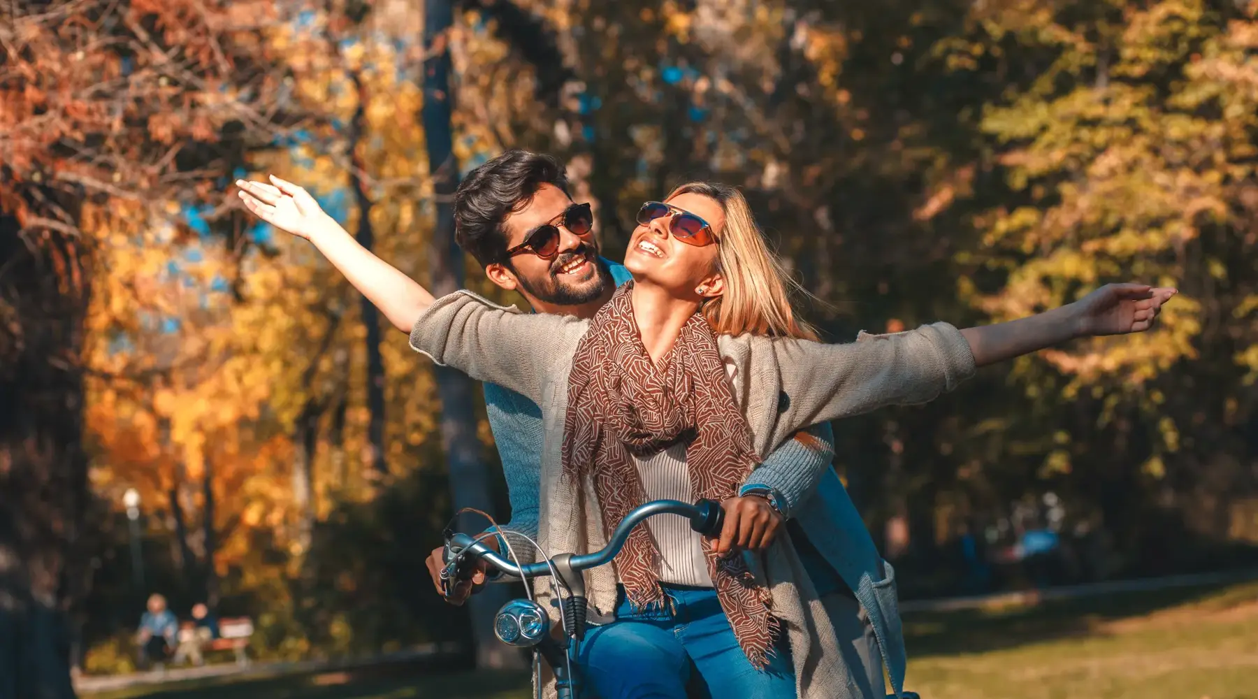 Two people ride a bicycle together through an autumn landscape and extend their arms. The scene conveys a sense of freedom, movement, and leisure activities in nature