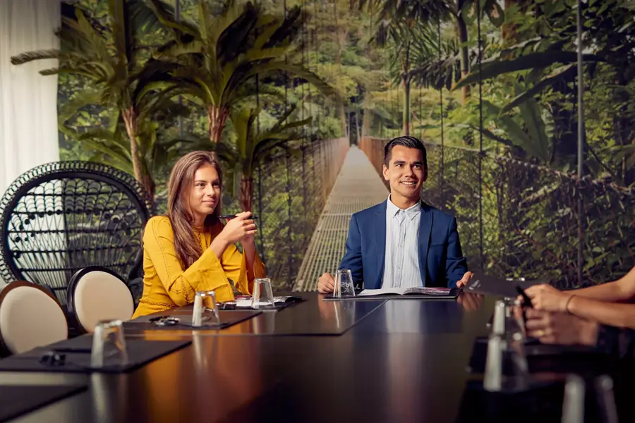Twee mensen zitten aan een vergadertafel in een tropisch ingerichte zaal met een jungle-achtergrond.