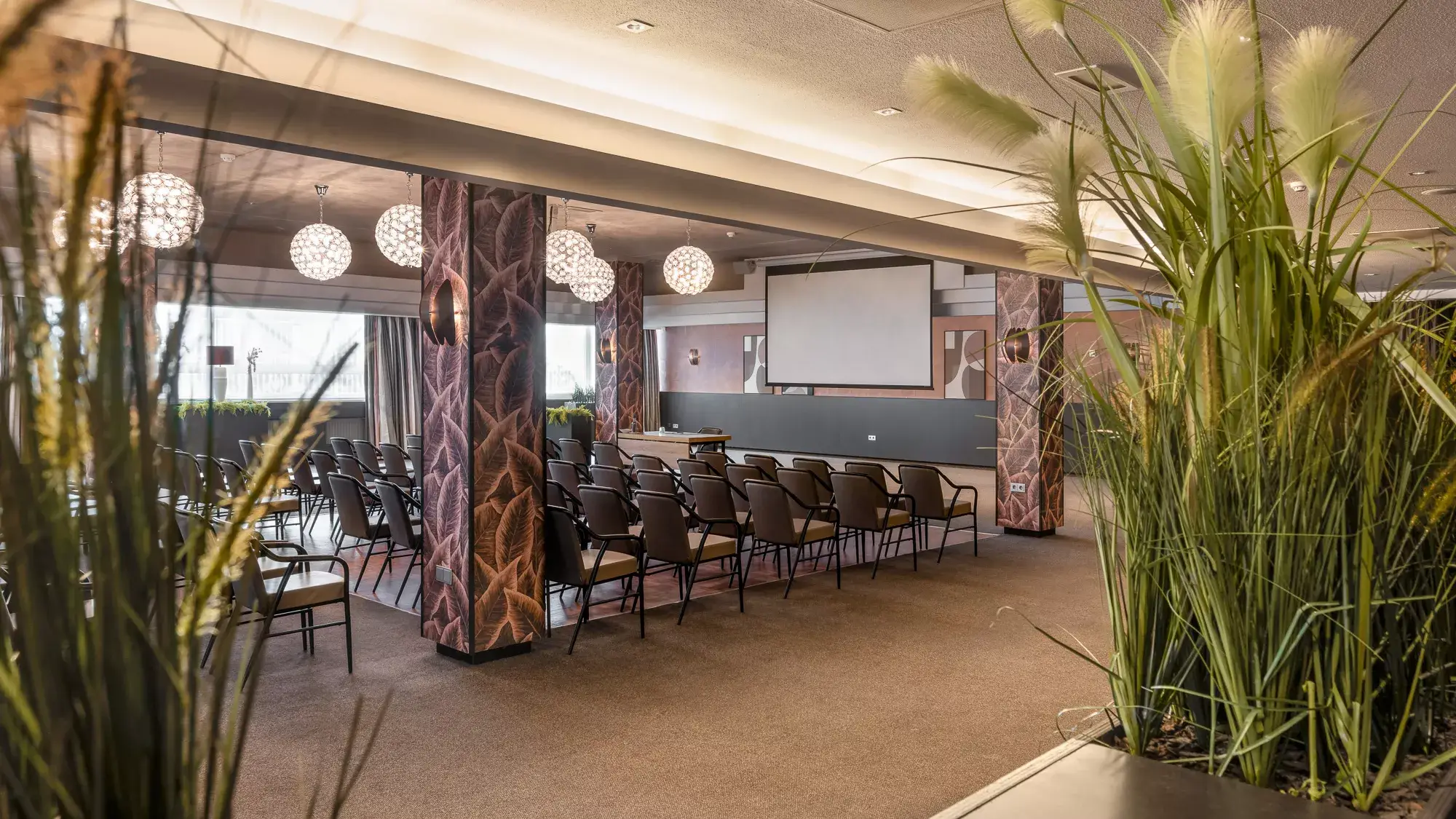 Sfeervolle doorkijk in de Valkenzaal langs groene beplanting, met zicht op de rijen stoelen en de presentatieruimte.