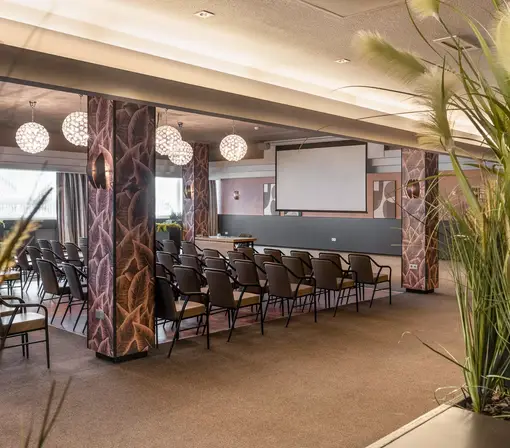 Sfeervolle doorkijk in de Valkenzaal langs groene beplanting, met zicht op de rijen stoelen en de presentatieruimte.