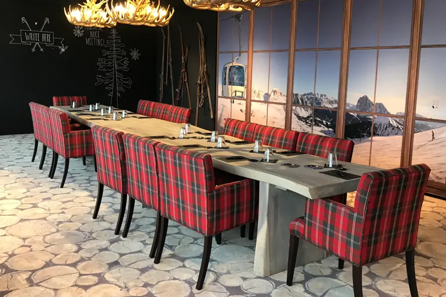 Een vergaderruimte in chaletstijl met een lange houten tafel, rode geruite stoelen en een wanddecoratie met besneeuwde bergen.