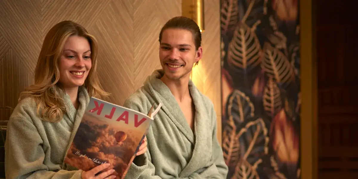 Zwei Personen sitzen entspannt im Wellnessbereich des Van der Valk Hotel Hildesheim, tragen Bademäntel und blättern gemeinsam in einem Valk Magazin. Die warme Beleuchtung und die ruhige Umgebung vermitteln eine entspannte, erholsame Atmosphäre.