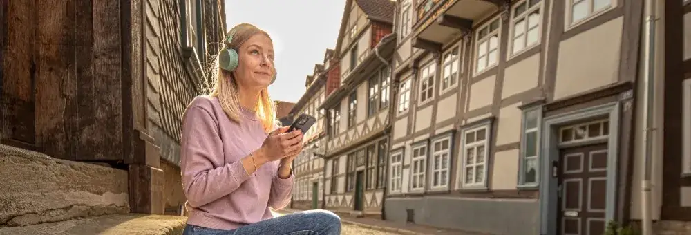 Lauschtour: Frau mit Kopfhörern sitzt in einer Altstadtstraße mit Fachwerkhäusern und nutzt ein mobiles Gerät, vermutlich für eine Audiotour.