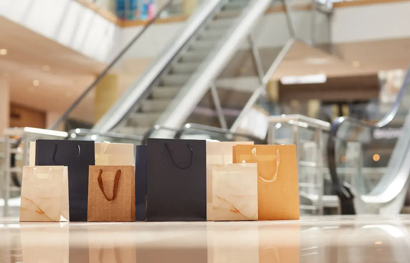 Verschillende boodschappentassen in verschillende kleuren staan op de vloer van een modern winkelcentrum. Op de wazige achtergrond zijn een roltrap en heldere binnenarchitectuur te zien, waardoor het beeld symbolisch staat voor winkelen en winkelmogelijkheden.