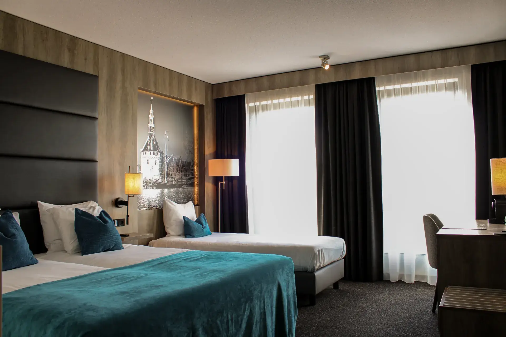 Spacious hotel room with a double bed, single bed, turquoise bedspread, blue decorative pillows, large windows, desk, and a black-and-white photo of the Hoofdtoren in Hoorn.