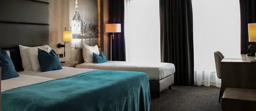Spacious hotel room with a double bed, single bed, turquoise bedspread, blue decorative pillows, large windows, desk, and a black-and-white photo of the Hoofdtoren in Hoorn.