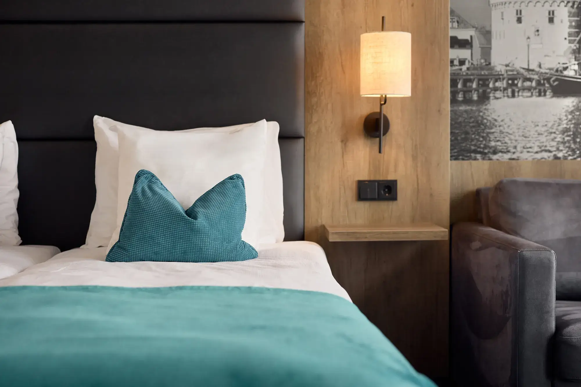 Detailansicht eines Hotelzimmers mit Bett, türkisfarbener Tagesdecke und Zierkissen, Holzvertäfelung an der Wand, Wandlampe, Steckdose neben dem Bett und grauem Sofa.