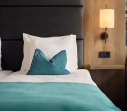 Detailansicht eines Hotelzimmers mit Bett, türkisfarbener Tagesdecke und Zierkissen, Holzvertäfelung an der Wand, Wandlampe, Steckdose neben dem Bett und grauem Sofa.