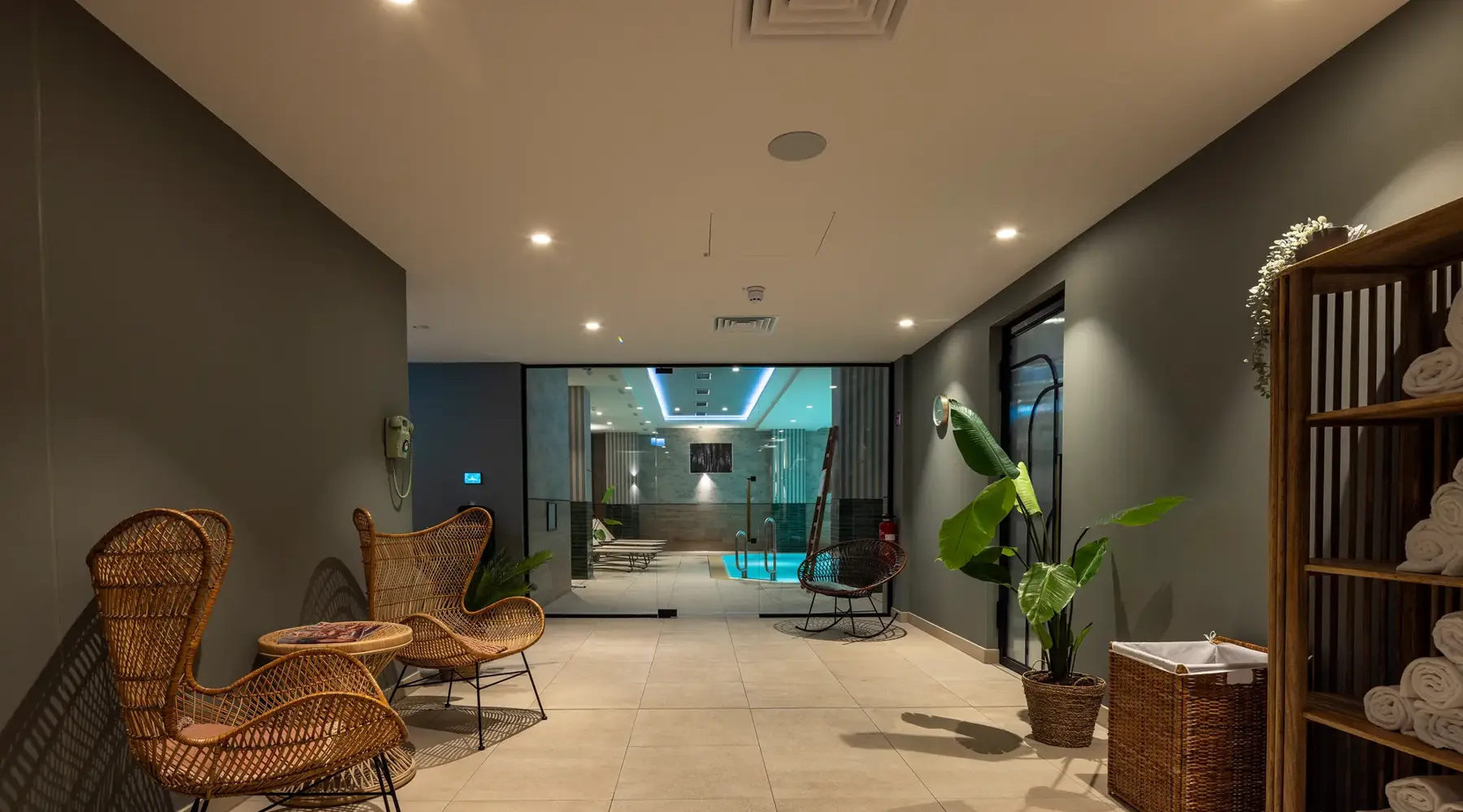 Quiet wellness area in the Van der Valk Hotel Hildesheim with tiled floors, plants, and wicker chairs. In the foreground, there are seating options and a shelf with towels, while in the background, the pool area is visible through a glass wall. The design looks modern, clean, and inviting.