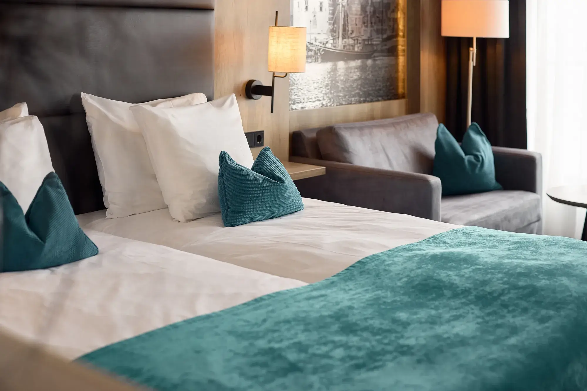 Detailansicht eines Hotelzimmers mit Doppelbett, türkisfarbener Tagesdecke und Zierkissen, grauem Sofa, Wandlampe und Steckdose neben dem Bett.