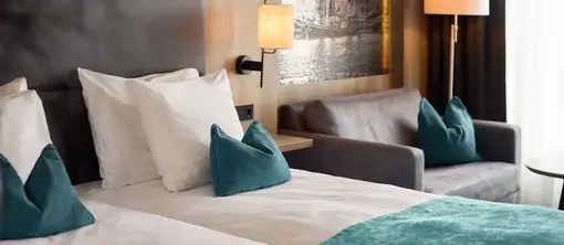 Detailansicht eines Hotelzimmers mit Doppelbett, türkisfarbener Tagesdecke und Zierkissen, grauem Sofa, Wandlampe und Steckdose neben dem Bett.