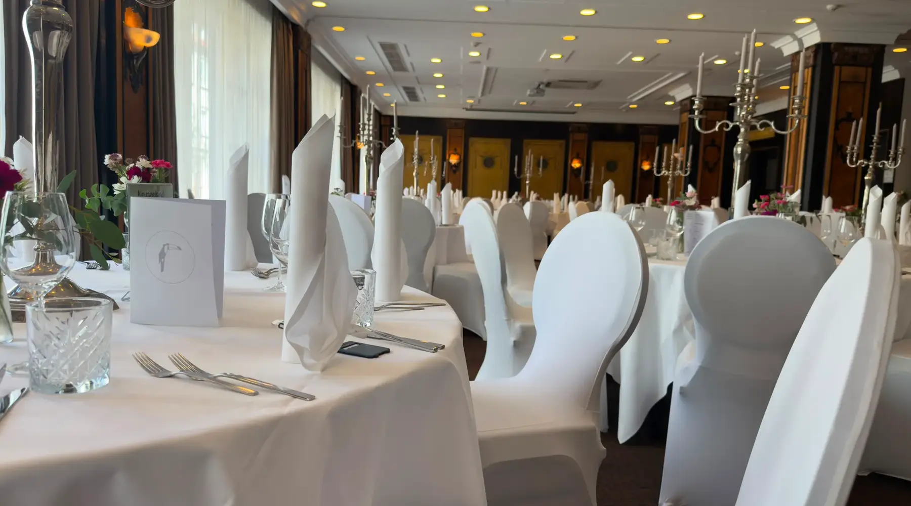 Feestelijk gedekte tafels met witte stoelhoezen, stoffen servetten en glazen in de Wollenweber-evenementenzaal van het Van der Valk Hotel Hildesheim.