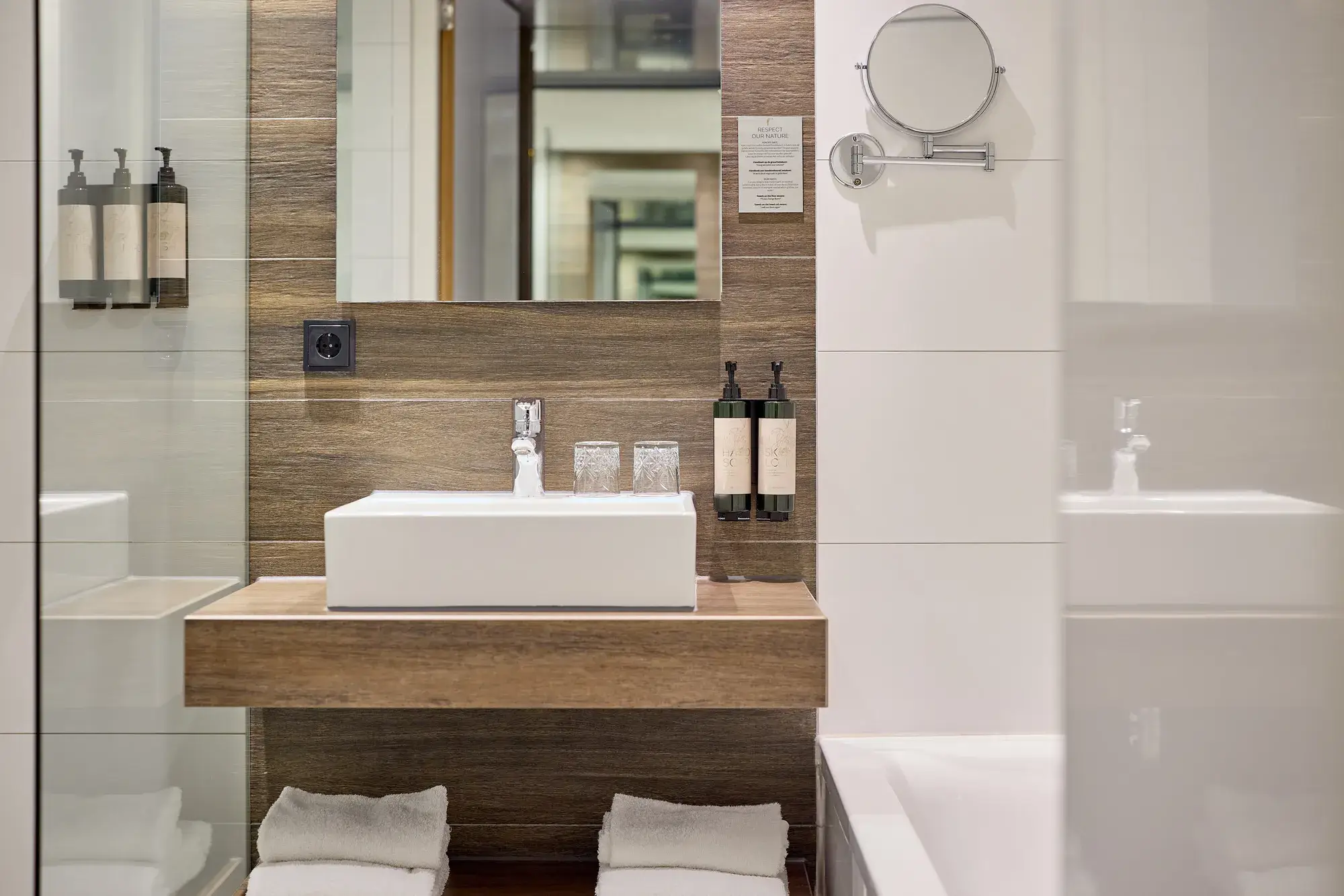 Modern hotel bathroom with white countertop washbasin, wooden vanity unit, large mirror, make-up mirror, toiletries, and towels.
