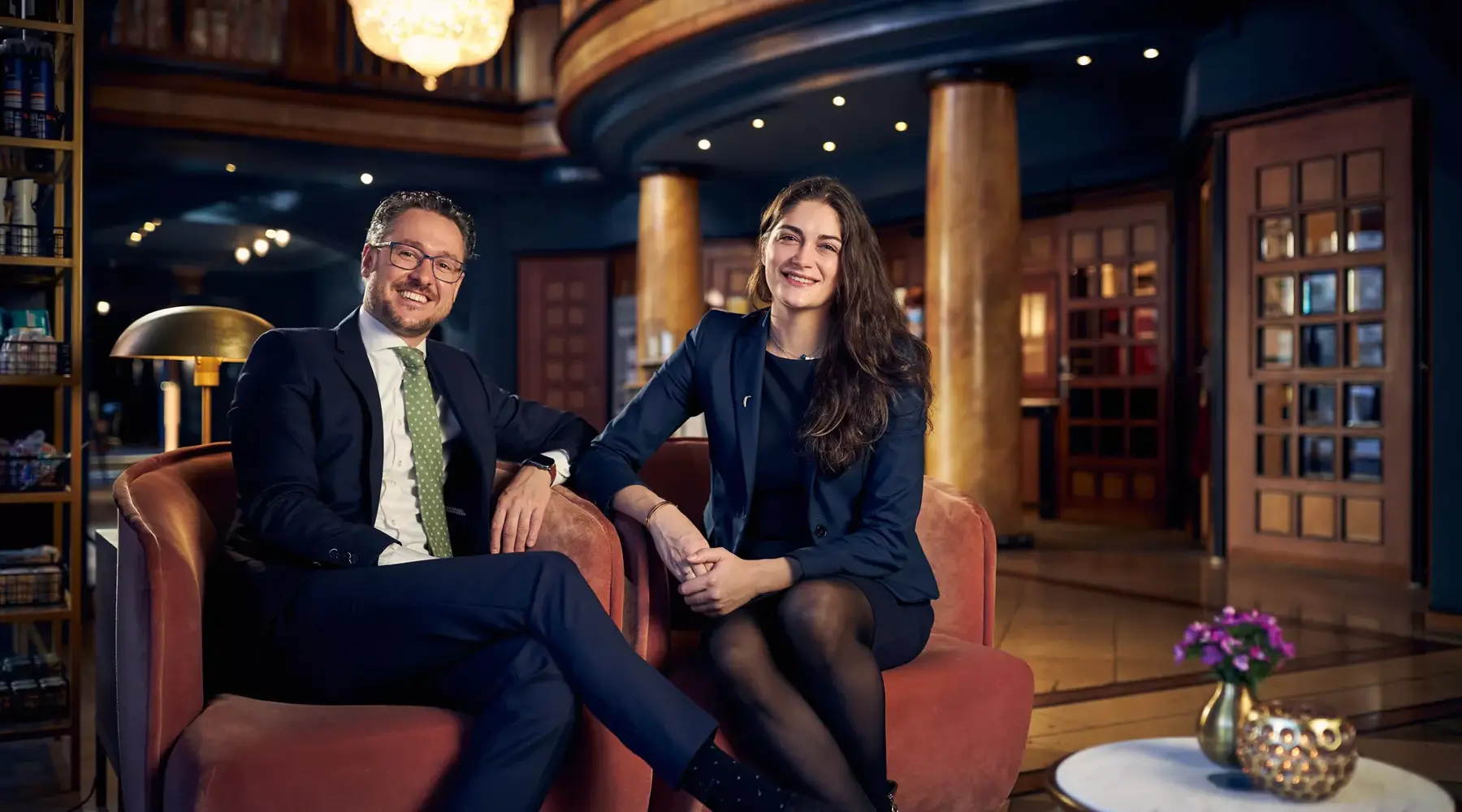 Het management van het Van der Valk Hotel Hildesheim heet u van harte welkom en staat voor kwaliteit, gastvrijheid en persoonlijke service. De twee zitten in de stijlvol vormgegeven lobby van het hotel; op de achtergrond zijn de open hotelhal en de receptie te zien. De warme verlichting en de hoogwaardige inrichting benadrukken de uitnodigende sfeer van het huis.