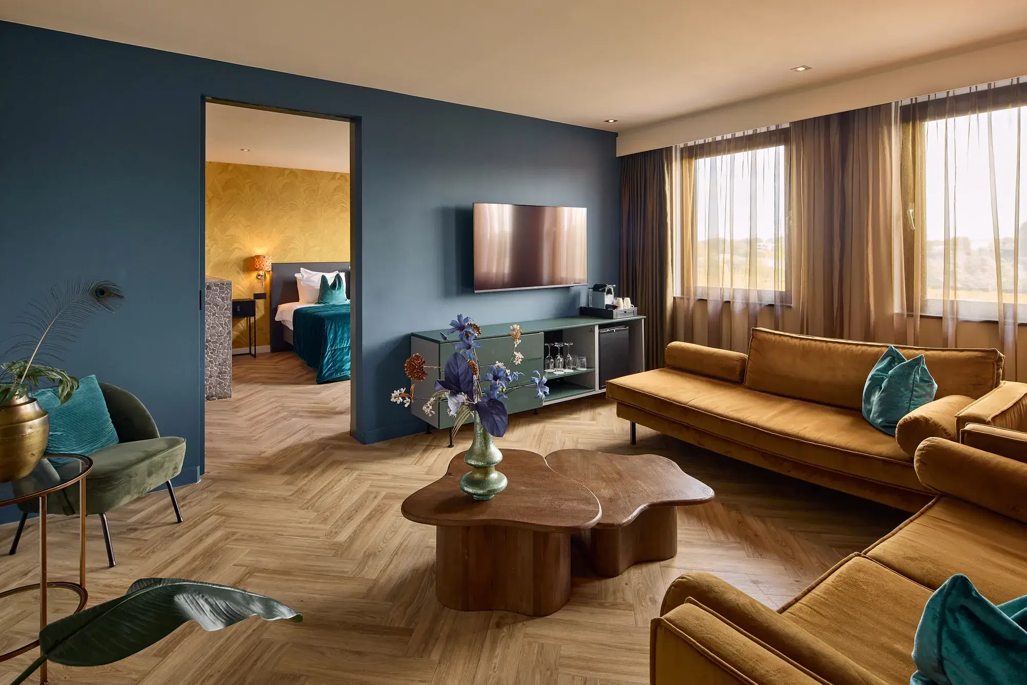 Luxe hotelsuite met ruime zithoek, okergele banken, groene fauteuil, houten salontafel, televisie, visgraatvloer en doorgang naar het slaapgedeelte.