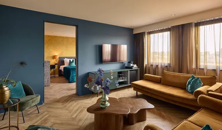 Luxe hotelsuite met ruime zithoek, okergele banken, groene fauteuil, houten salontafel, televisie, visgraatvloer en doorgang naar het slaapgedeelte.