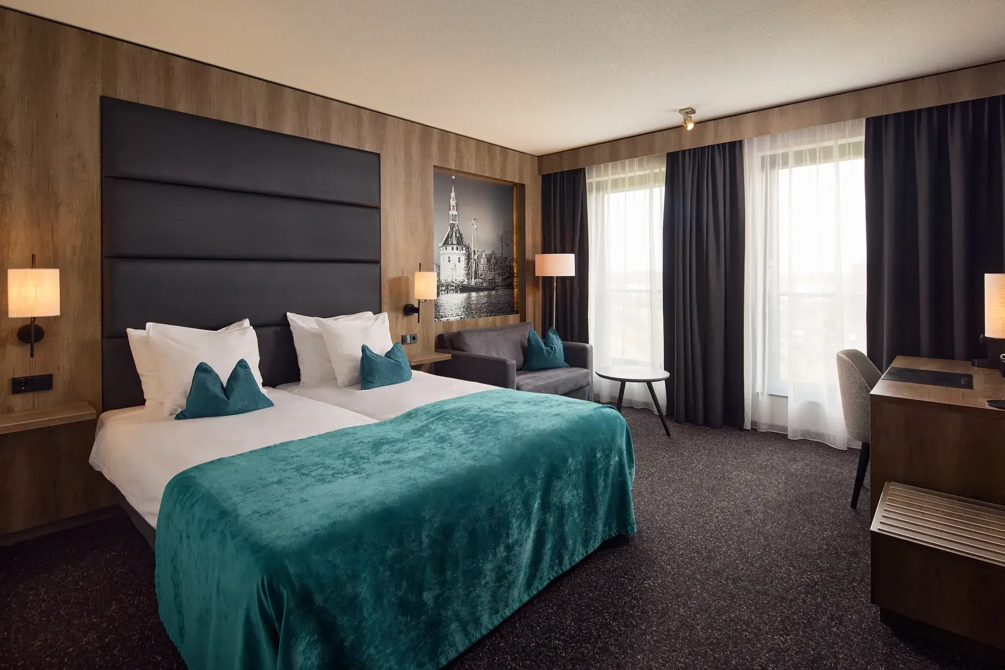 Geräumiges Hotelzimmer mit Doppelbett, türkisfarbener Tagesdecke, Sitzecke mit Sofa, Schreibtisch, großen Fenstern und einem Schwarz-Weiß-Foto des Hauptturms in Hoorn.