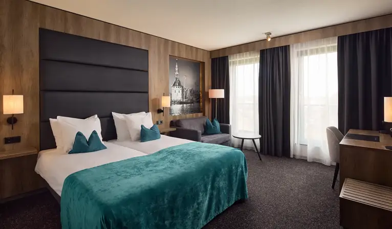 Geräumiges Hotelzimmer mit Doppelbett, türkisfarbener Tagesdecke, Sitzecke mit Sofa, Schreibtisch, großen Fenstern und einem Schwarz-Weiß-Foto des Hauptturms in Hoorn.