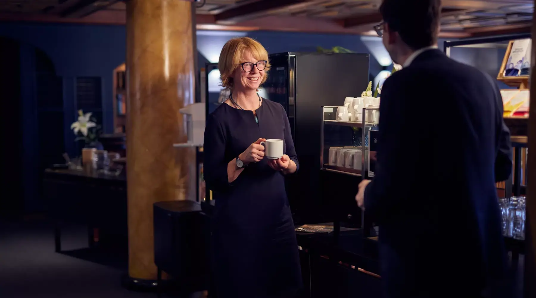 Eine Frau steht im Van der Valk Hotel Hildesheim in einem stilvollen Veranstaltungsbereich und hält eine Tasse Kaffee in der Hand, während sie sich mit einer weiteren Person unterhält. Im Hintergrund sind eine Kaffeemaschine, Tassen und Gläser zu sehen. Die Szene vermittelt eine entspannte Pause während einer Tagung oder eines Meetings.