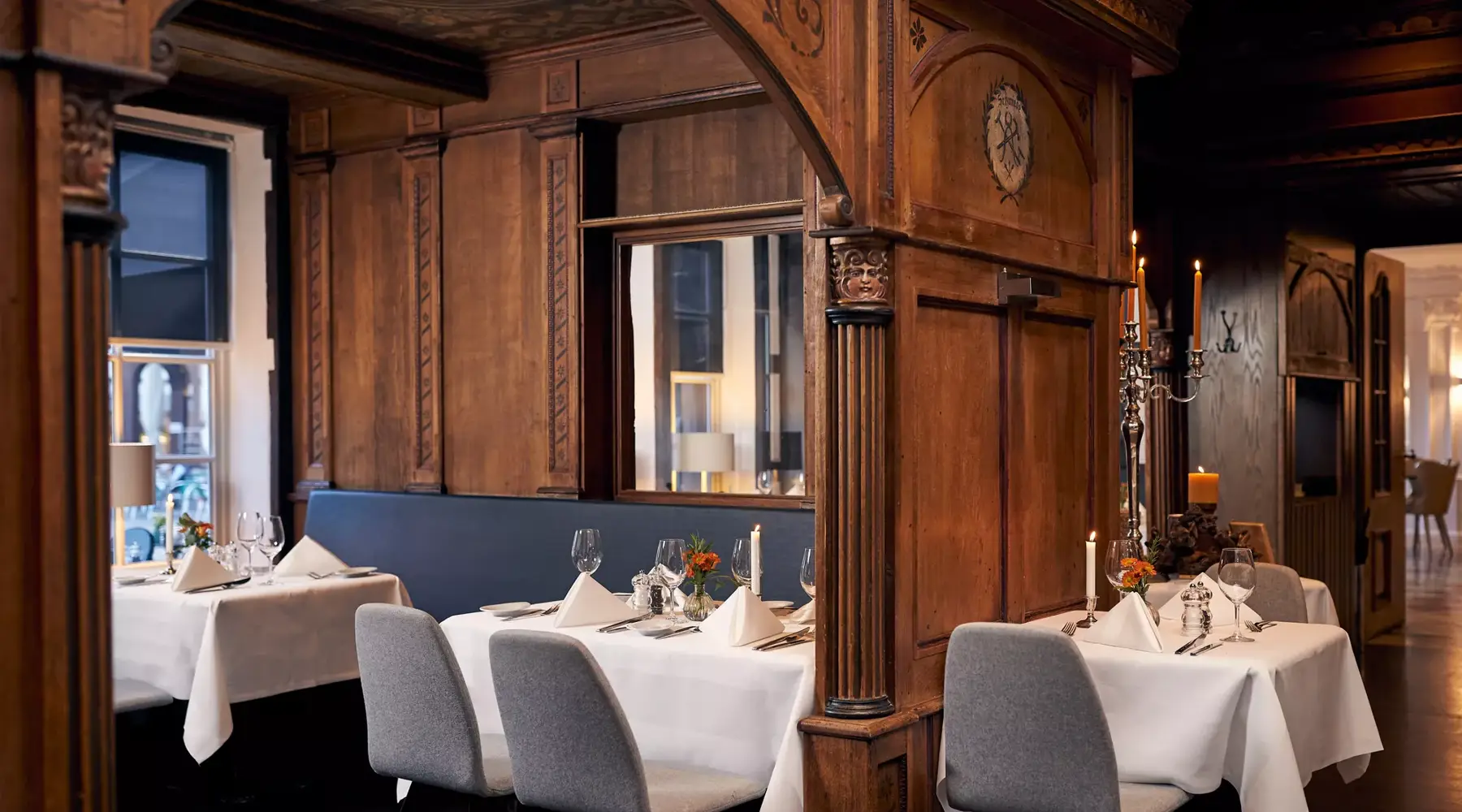 Stilvolle Restaurantnische im Van der Valk Hotel Hildesheim mit warm vertäfelten Holzwänden und dekorativen Schnitzdetails. Mehrere Tische sind mit weißen Tischdecken, gefalteten Servietten, Gläsern und Kerzen elegant eingedeckt, umgeben von gepolsterten Stühlen. Gedämpftes Licht und klassische Elemente wie Kerzenständer und Wandverzierungen schaffen eine ruhige, hochwertige und intime Atmosphäre.