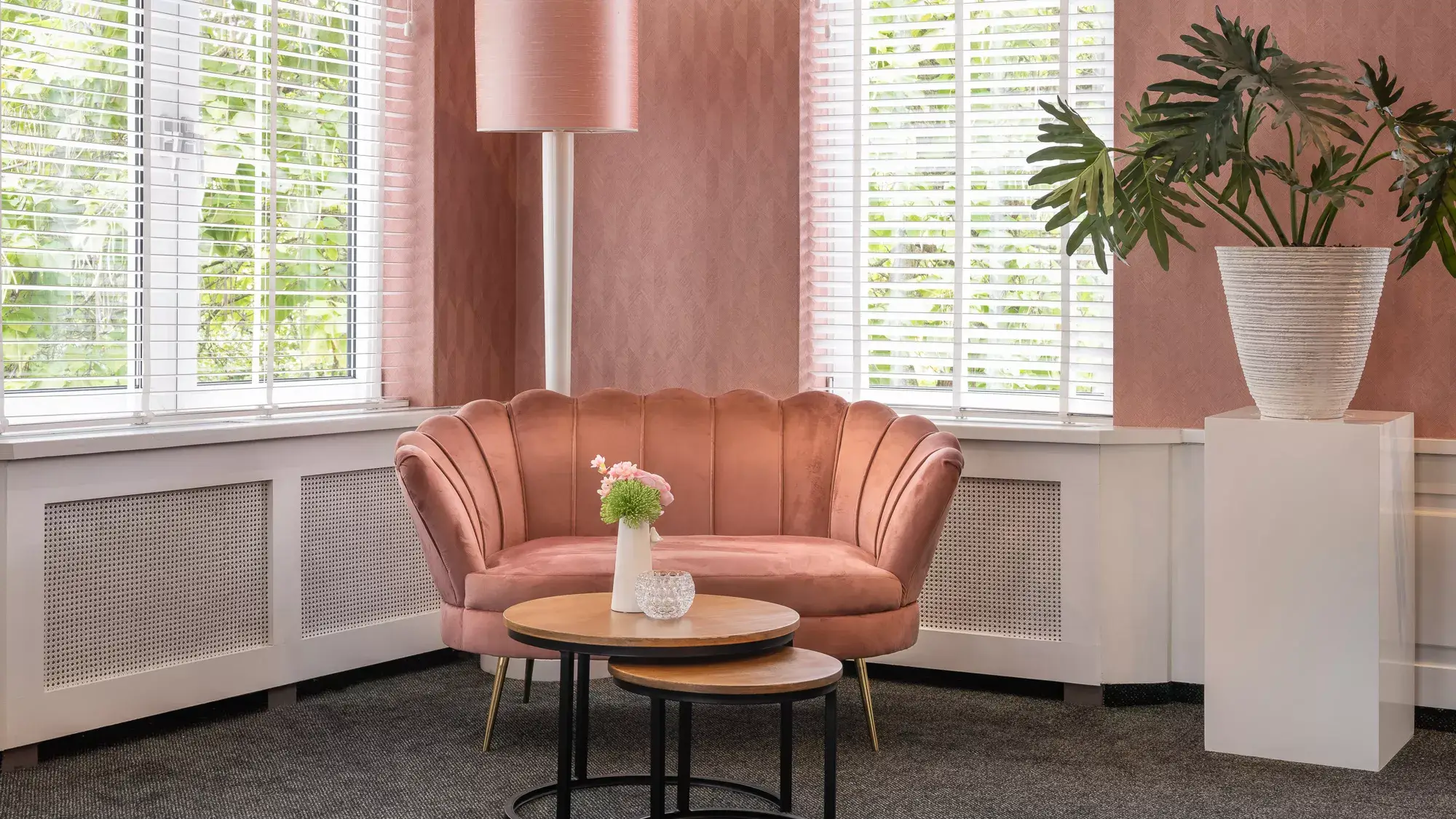 Detail van een comfortabele zithoek in de ruimte, passend ingericht met een roze bankje, tafeltjes en plantendecoratie.