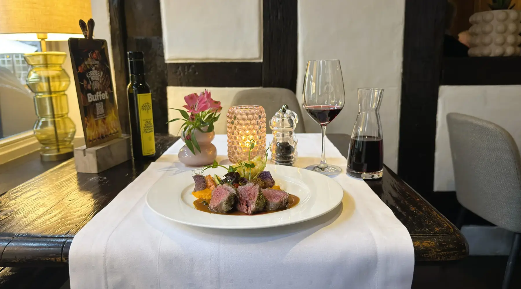 An elegantly plated meat dish with vegetables and sauce on a white tablecloth in a restaurant, served with a glass of red wine, a carafe, candlelight, and floral decoration.