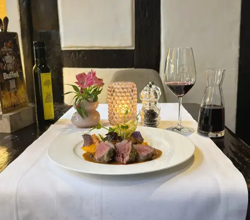 Een stijlvol opgemaakt vleesgerecht met groenten en saus op een wit gedekte restauranttafel, geserveerd met een glas rode wijn, een karaf, kaarslicht en een bloemendecoratie.