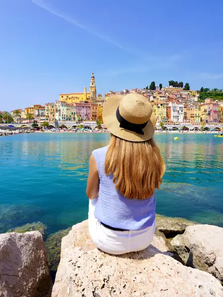 Tourist with a sunhat on a rock, looking at a colorful seaside town in southern France