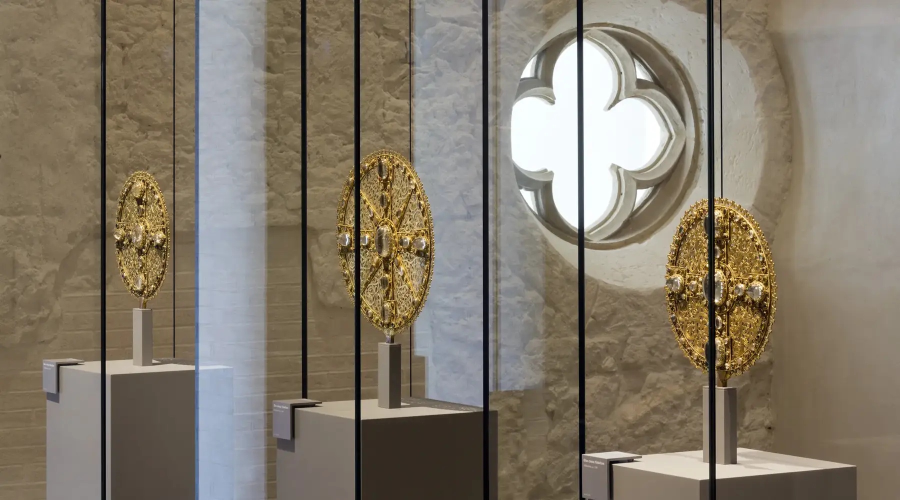 Several artistically decorated, golden liturgical objects are displayed in a modern glass case. They stand on simple pedestals in front of a light stone wall with a round window featuring a cross-shaped opening. The interplay of historical sacral art and modern presentation creates a calm, museum-like atmosphere.