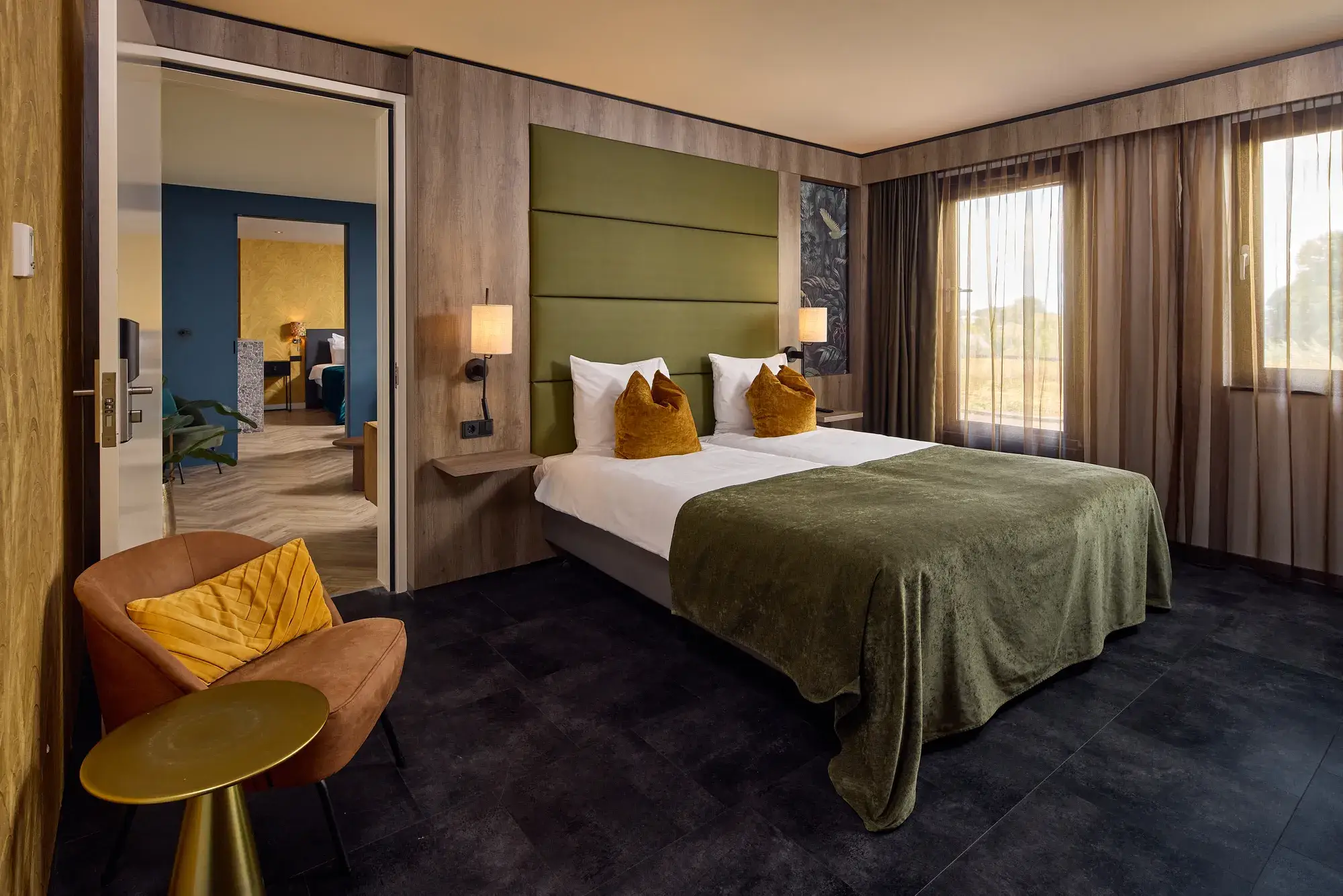 Ruime hotelkamer met tweepersoonsbed, groene bedsprei, okergele sierkussens, groene wandbekleding, fauteuil met bijzettafel en doorgang naar een aangrenzende kamer.