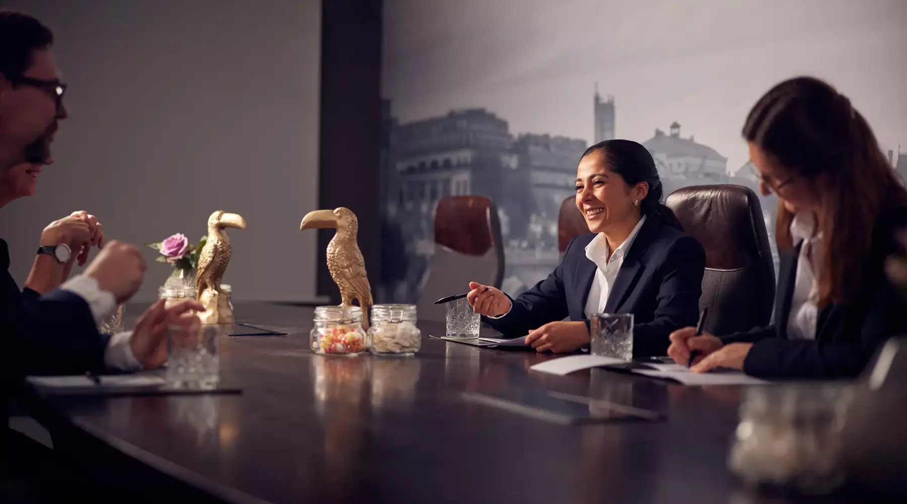 Several people are sitting at a conference table in the Van der Valk Hotel Hildesheim and having a conversation. A woman in a business outfit smiles and holds a pen in her hand, with notes and glasses in front of her. On the table, there are decorative elements like small glasses and figures that give the room a stylish, professional atmosphere.