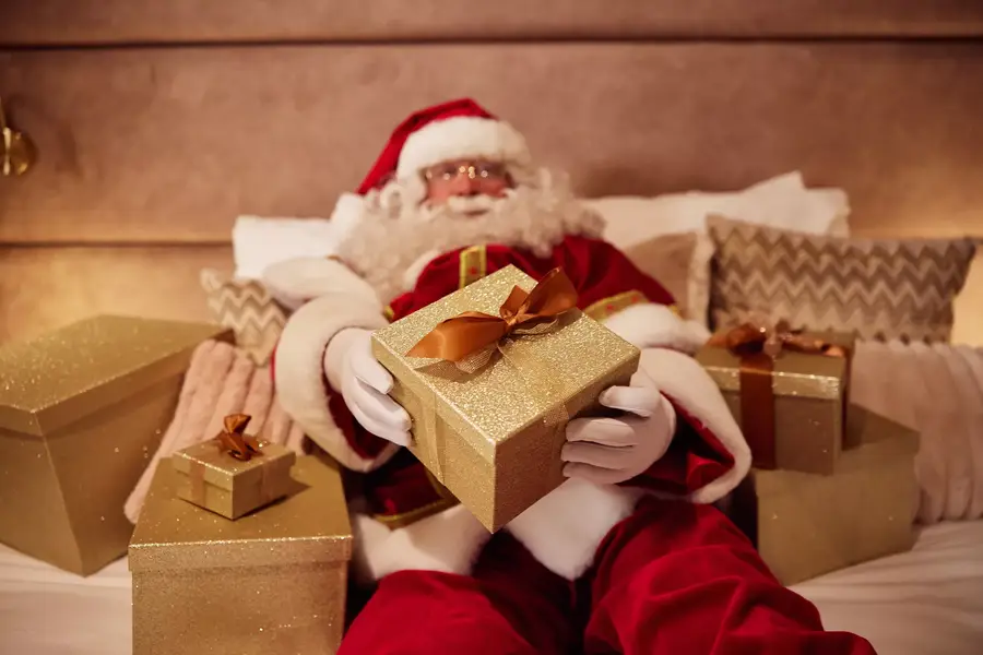 De Kerstman ontspant met cadeaus op een comfortabel hotelbed, als speels sfeerbeeld voor een zorgeloze kerstovernachting.