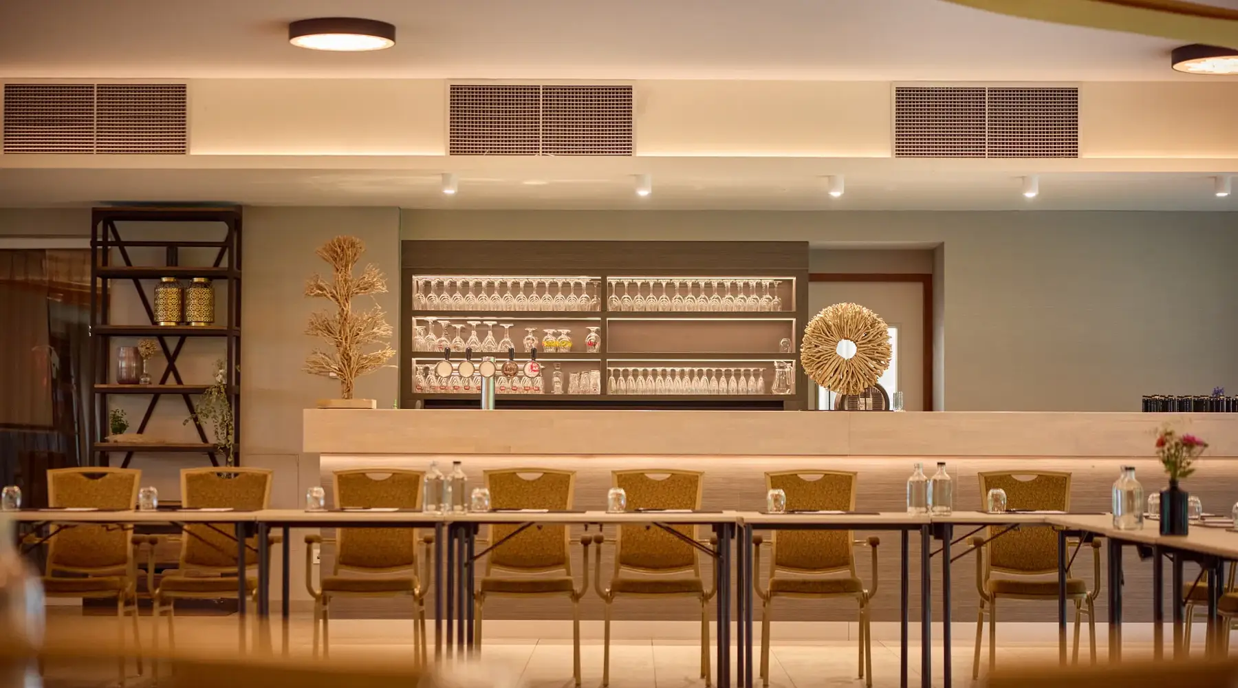 Een vergaderzaal in Hotel Dennenhof met lichte beige kleuren. Een tafelopstelling in U-vorm met stoelen en op de achtergrond een bar met glazen en decoratie.